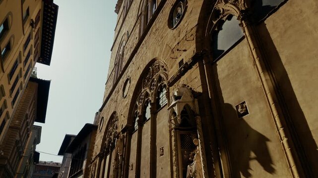 A carved saint stands in a canopied Gothic tabernacle on the tall stone flank of Orsanmichele in Florence, Tuscany. Street level low angle, slow forward drift.