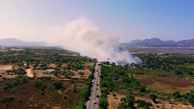Cars keep moving along a country road past scattered houses as wildfire smoke drifts low across the valley near Olbia, Sardinia, Italy. Aerial view, forward tracking shot.