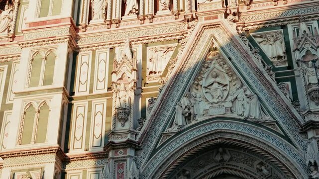 A sculpted tympanum sits above one of the Duomo portals, framed by the green and pink marble banding of the cathedral front in Florence, Tuscany. Street level close up, diagonal drift.