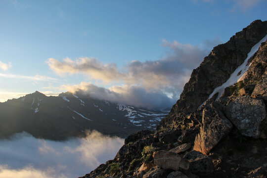 Dramatic mountain landscape near Anchorage, Alaska, with rugged rocky peaks, lingering snow and low clouds below in the valley. Scenic outdoor adventure and travel concept.