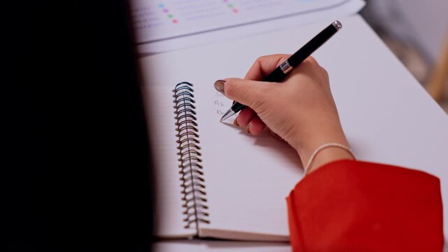 Hand Writing in Spiral Notebook with Black Pen, Red Sleeve, Bracelet, Desk Study Session, Education Planning, Personal Organization, Focused Note Taking and Creative Learning Practice