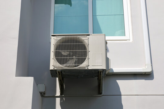 Air conditioner outdoor condenser unit installed on the wall of a modern residential house beside a window.