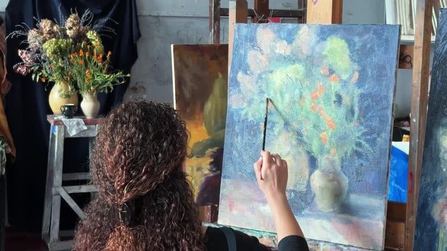 Teenage girl in college painting class, art studio, art workshop. She paints still life on canvas with oil paints, holds brush, palette.