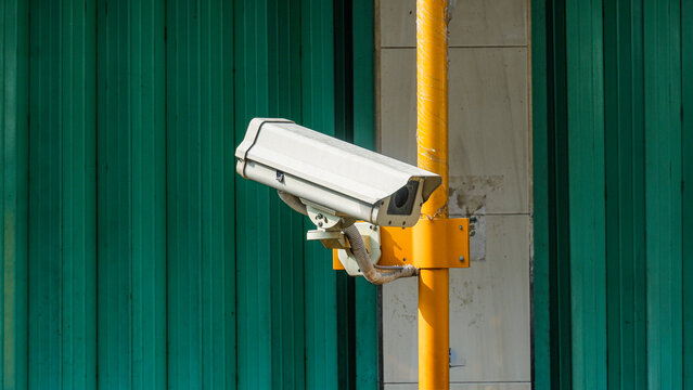 Outdoor CCTV security camera mounted on a bright yellow post against a green corrugated metal wall background.