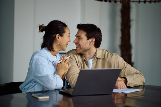 Happy multiracial couple talking while working on laptop at home.