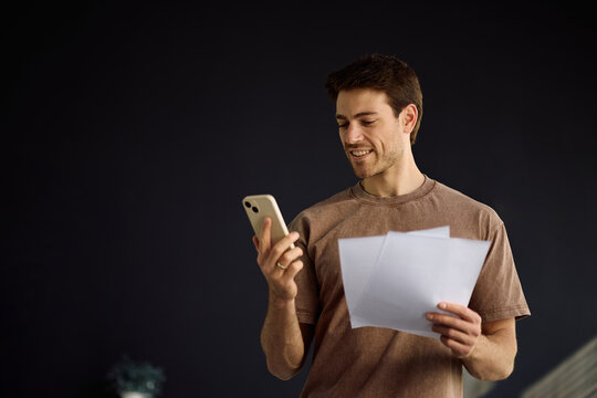 Happy man using cell phone app while going through financial reports at home.