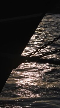 Silhouetted boat hull and chain with golden sunlight reflecting on calm, shimmering water. Serene and peaceful vertical footage.
