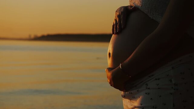 Close-up of pregnant woman gently holding her baby bump by calm water at sunset, capturing motherhood, pregnancy, anticipation and maternal care in warm evening light.