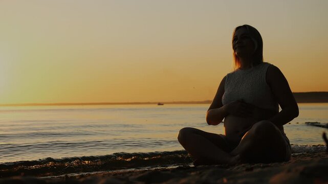 Pregnant woman sits by calm water at sunset holding her baby bump and looking into the distance, symbolizing motherhood, anticipation, maternity and peaceful pregnancy.