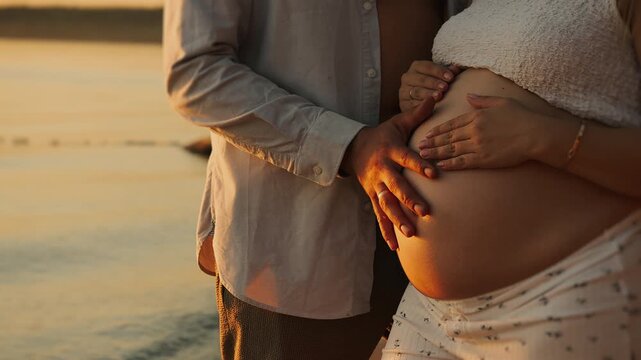Expecting couple gently holding pregnant belly together by the water at sunset, symbolizing pregnancy, parenthood, family bonding and anticipation of a baby.