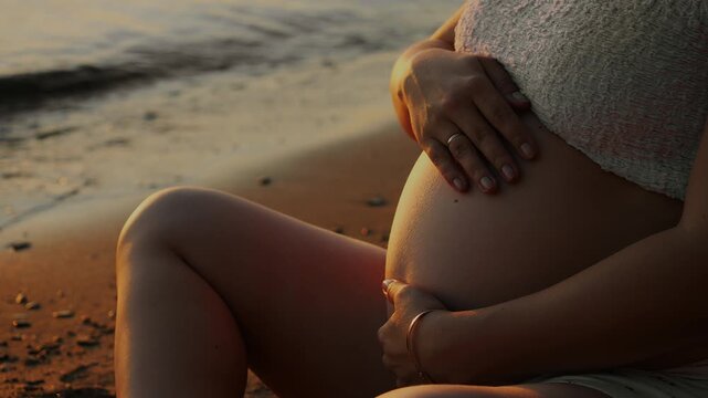 Close-up of pregnant woman sitting by the water and gently holding her baby bump at sunset, representing motherhood, maternity, prenatal wellbeing and anticipation.