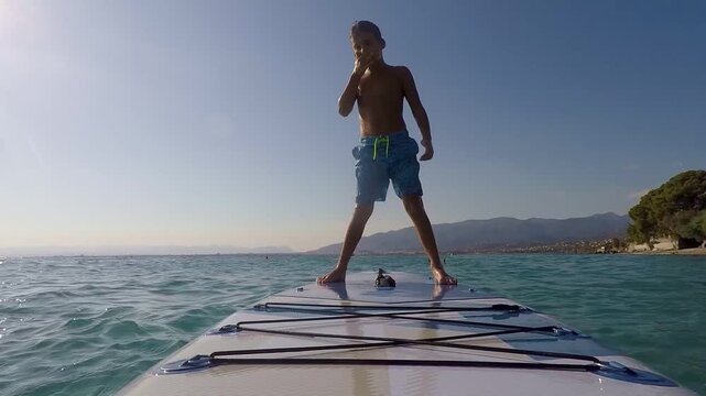 A young boy balances on a paddleboard in the ocean waves on a sunny day
