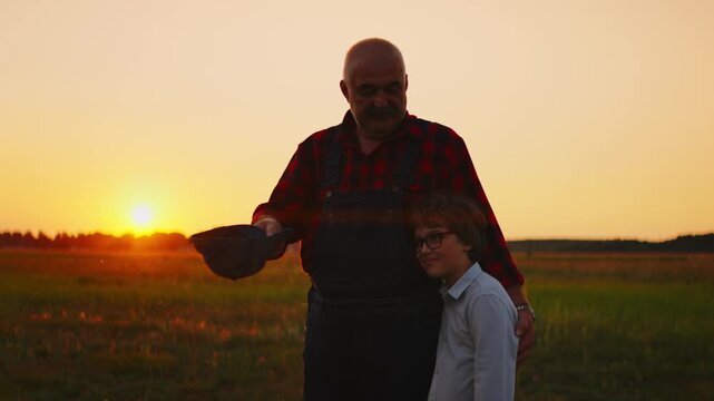 Grandfather and grandson standing together in a peaceful rural field at sunset, sharing a quiet moment of family connection, love, and generational farming heritage