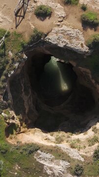 Vertical aerial rising shot over the Portim&atilde;o coast, highlighting the rock formations, coastal caves, vegetation, and beaches around Gruta das Baratas.