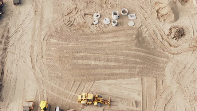 Abstract overhead aerial of pale swept sand with sweeping track marks, small yellow machines and staged materials arranged across the flat working surface.