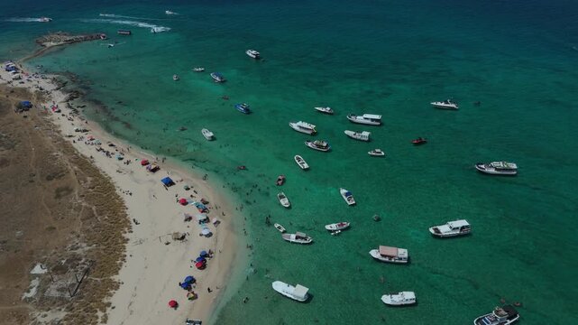 An aerial drone shot pans across a crowded beach with clear turquoise water, anchored boats, and people swimming at Rabbit Island in Tripoli, Lebanon.