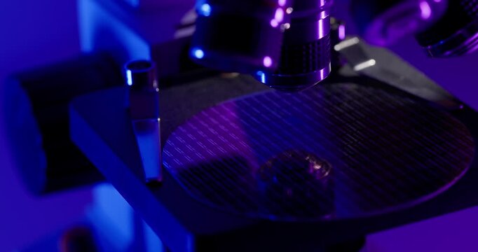Slow motion microscope inspecting silicon wafer with microchip under purple and blue lab lighting on research bench.