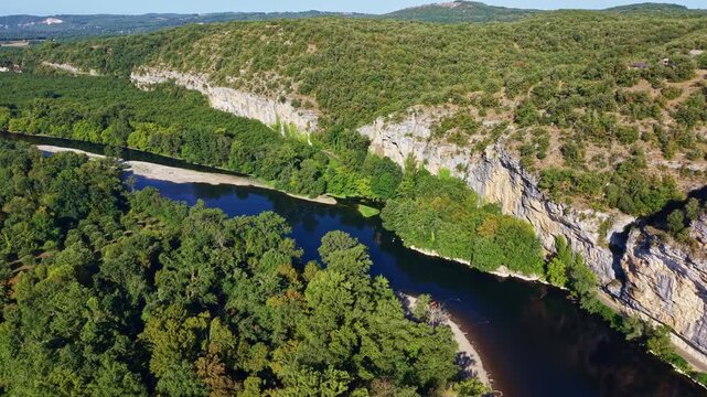 Dordogne River flows below the Roc des Monges limestone escarpment near Saint-Sozy, Lot, Occitanie, France, with exposed riverbank sediment and dense riparian woodland, drone forward flyover
