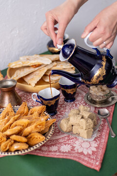 Middle Eastern Coffee and Sweets Spread. Hand pours coffee from a cobalt blue vintage decanter pot in cup. Sweet food on table honey baklava and halva on a patterned red tablecloth.