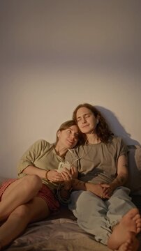 Vertical video of young Caucasian couple sharing wired earphones and listening to music from smartphone while relaxing together indoors. Perfect for streaming app promo and mobile lifestyle ads