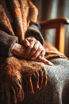 Hands of an elderly woman rest on her lap covered by a warm shawl while sunlight streams through the window, creating a bright atmosphere in her home