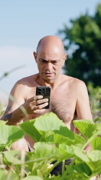 Mature shirtless farmer sitting in his urban garden while using a mobile phone. A man checking his zucchini plants with modern technology. Vertical