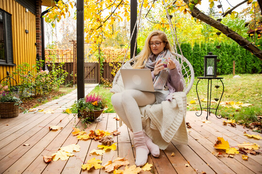 A mid adult woman works remotely on a laptop while drinking tea in a hanging swing chair on an autumn patio.