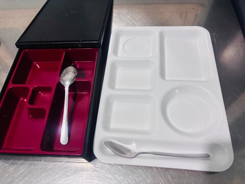 Empty lunch trays, including a compartmentalized black and red bento box and a white cafeteria tray with metal spoons on a metal table.