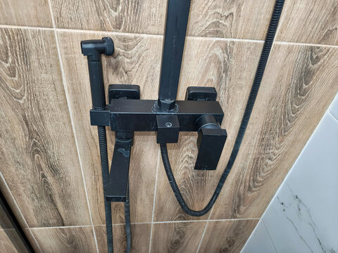 Modern black square uncleaned shower faucet installed on brown wood effect ceramic wall tile. Bathroom interior plumbing fixture with handheld sprayer for daily hygiene