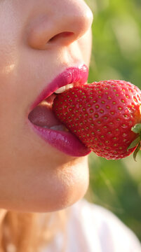 A captivating close-up features a woman's mouth with bright pink lipstick sensually biting into a fresh, ripe red strawberry. This image evokes the joy of healthy eating, summer freshness, and the...