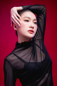 An artistic portrait with an arm curved gracefully overhead, gaze lifted contemplatively, elongated neckline and sheer black fabric creating a flowing silhouette against deep red.