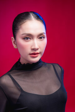 A poised beauty portrait with a direct, symmetrical gaze, sleek center-parted hair and sheer black attire against a rich red backdrop, exuding calm confidence.