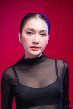 A poised beauty portrait with a direct, symmetrical gaze, sleek center-parted hair and sheer black attire against a rich red backdrop, exuding calm confidence.