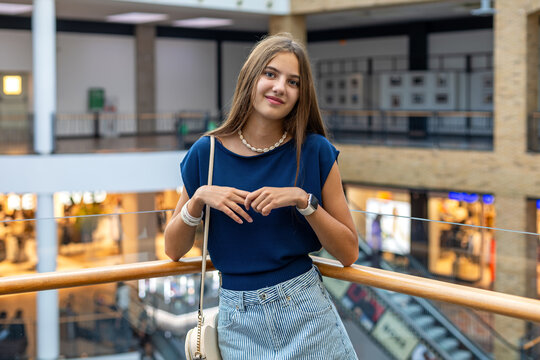 Young Woman Standing in Modern Shopping Mall. Young woman smiles while standing by a railing inside a modern shopping mall with stores and an escalator behind.