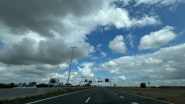 Wide highway stretching beneath dramatic storm clouds with blue sky openings, light traffic, roadside signage, tall lamp posts, and open landscape creating strong travel and weather contrast