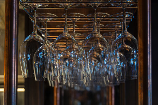 Hanging Wine Glasses on Rack in Warm Bar Interior. Rows of empty wine glasses hang upside down from a metal rack, reflecting warm and cool light inside a softly lit bar interior.