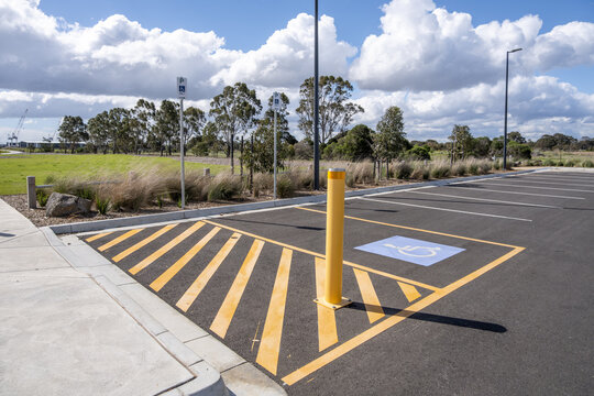 An empty outdoor public car park with a designated accessible parking space marked with a wheelchair symbol, yellow hazard diagonal lines, a safety bollard, and concrete curb ramping in Australia