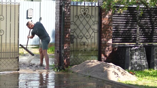 Man uses a heavy squeegee broom to forcefully push muddy floodwater out of his suburban private house driveway.
