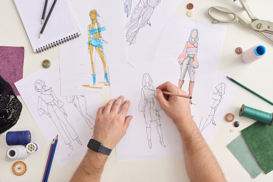 Fashion designer sketches garment ideas on paper at workspace. Hands draw figure drawings beside fabric swatches, buttons, thread spools, and scissors, showing focused creative work and careful