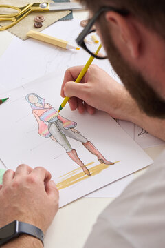 Designer draws clothing illustration with colored pencil on white paper. Fashion sketching continues beside scissors, buttons, and fabric swatches, while focused artist refines stylish outfit.