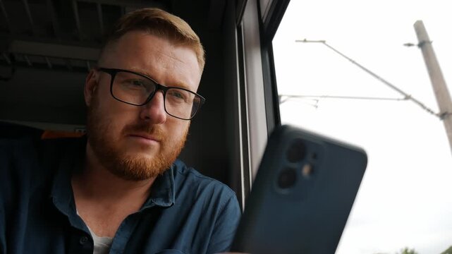 Young bearded man in glasses looking at smartphone screen while sitting by window inside moving train. Focused passenger browsing internet, texting, and commuting during railway journey.