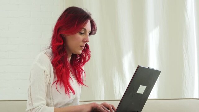 Young Woman with Vibrant Red Hair Working on a Laptop at Home in a Bright Living Room