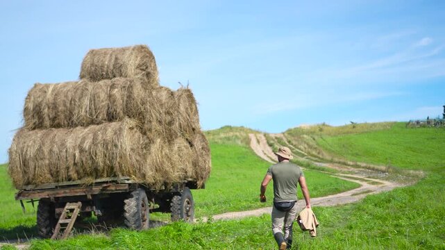 Highly engaging commercial ready wide shot of a male farmer walking away up a winding hill road, waving hand while passing by a massive loaded hay wagon.
