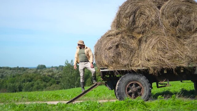 Highly engaging commercial ready medium shot of a handsome male farmer posing with confidence next to a large loaded hay wagon in a summer field.
