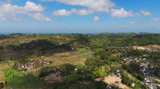 Red roofed village nestles among terraced rice fields in Lombok valley, forested hills rise toward distant coastline, scattered clouds drift above tropical countryside. Aerial drone wide shot