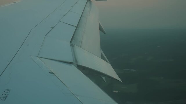 Airplane wing shaking during turbulence, flaps extended for landing approach over green landscape. Passenger aircraft window view of bumpy descent, technical wing mechanism work close up.