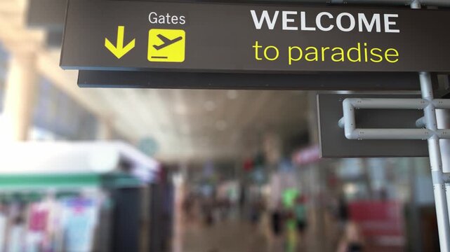 Close up view of an airport arrival sign with baggage claim directions and a Welcome to Paradise message, with a blurred background of passengers walking through the terminal hall