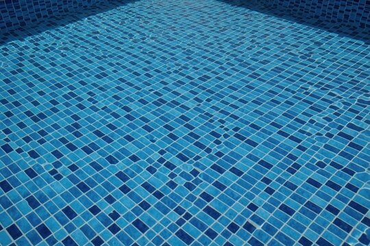 Blue mosaic tiles cover bottom of swimming pool. Clear water ripples over small square patterns. Summer vacation background features bright turquoise texture in luxury condominium resort. Surface