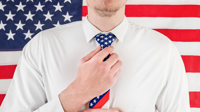 Man in a white shirt adjusts a patriotic necktie before an American flag