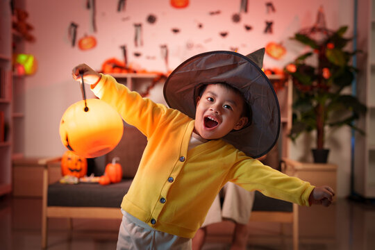 Asian child wearing witch hat holding glowing pumpkin lantern in decorated home for Halloween